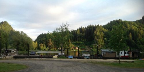 A serene lakeside view with boats docked, surrounded by lush green hills and trees under a cloudy sky.