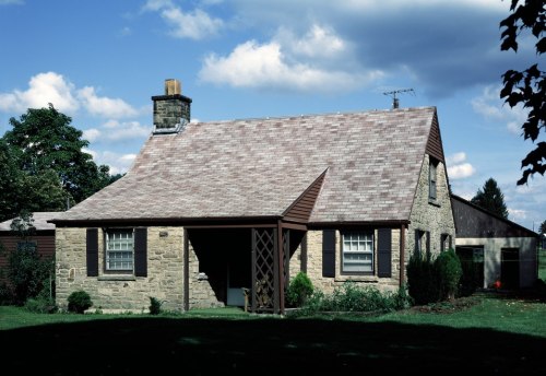 A quaint stone cottage with a sloped roof, surrounded by greenery and under a partly cloudy sky.