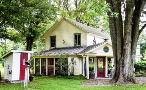 A charming two-story house with a sloped roof, surrounded by trees and a small shed nearby.