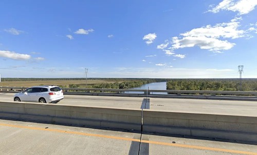 A car drives on a bridge overlooking a river and lush greenery under a clear blue sky.