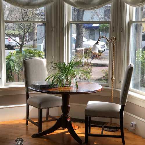 A cozy corner with a round wooden table, two chairs, and a potted plant, surrounded by large windows.