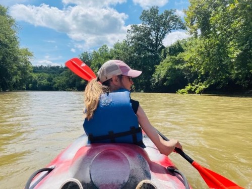 A person in a kayak paddles on a river surrounded by lush greenery under a bright blue sky.