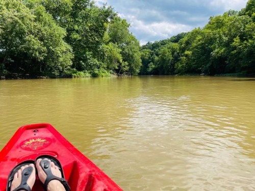 A person’s feet in sandals on a red kayak, floating on a calm, muddy river surrounded by lush green trees.