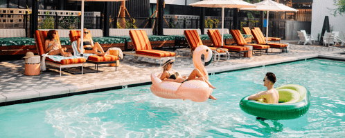 Two people relax in a pool on inflatable floats, surrounded by lounge chairs and umbrellas on a sunny day.