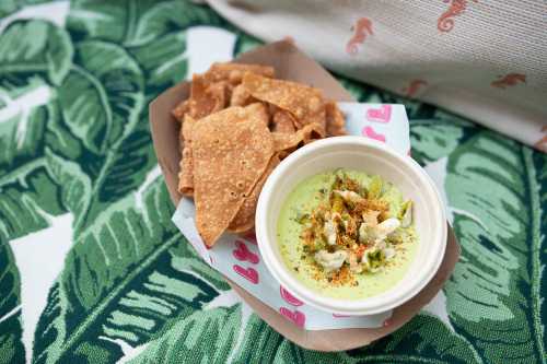 A bowl of green dip topped with spices, served with crispy tortilla chips on a leafy patterned tablecloth.