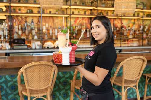 A smiling server holds a tray with colorful cocktails in a vibrant bar setting.