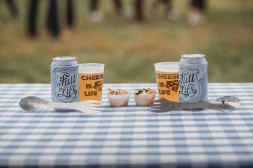 Two cans of beer and cups on a checkered tablecloth, with two small containers of food in the center.