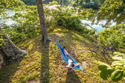 A person relaxes in a blue hammock by a serene river, surrounded by trees and bicycles nearby.