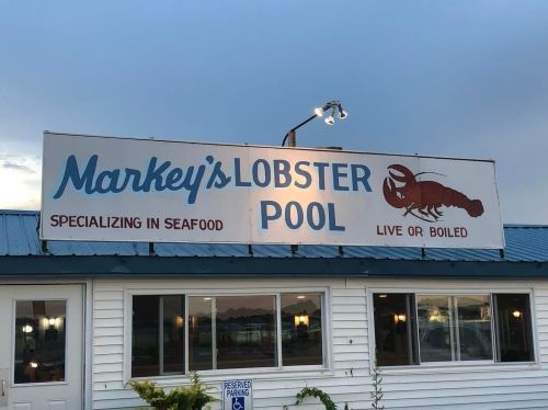 Sign for Markey's Lobster Pool, featuring a lobster graphic and text about seafood options, under a cloudy sky.