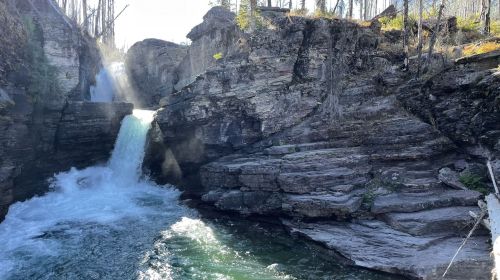 A serene waterfall cascades over rocky cliffs, surrounded by trees and sunlight, creating a tranquil natural scene.