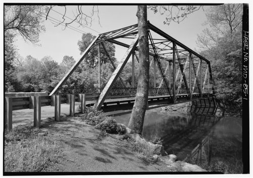 Black and white image of a metal truss bridge over a calm river, surrounded by trees and a gravel path.