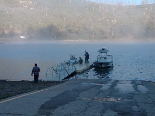 A misty lake scene with a dock, two boats, and people preparing equipment on the shore.