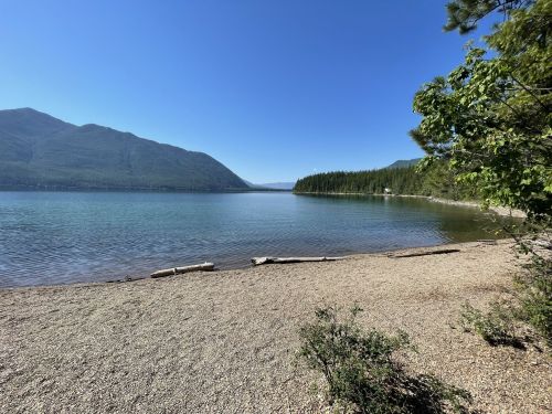 A serene lake surrounded by mountains and trees, with a sandy shore under a clear blue sky.
