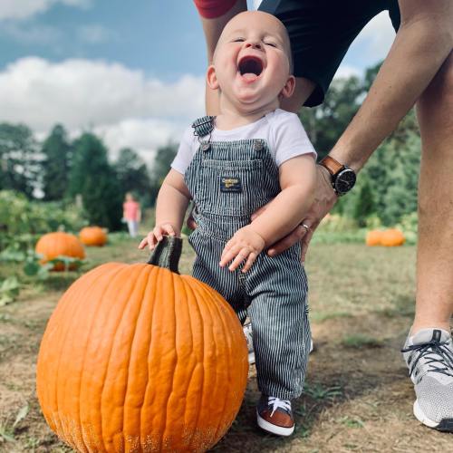 A joyful toddler in overalls laughs while standing next to a large pumpkin in a pumpkin patch.