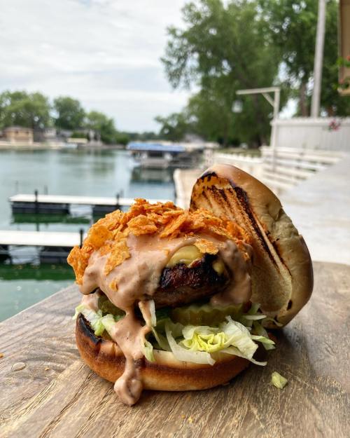 A close-up of a burger with lettuce, sauce, and crispy toppings, set on a wooden table by a lake with boats in the background.