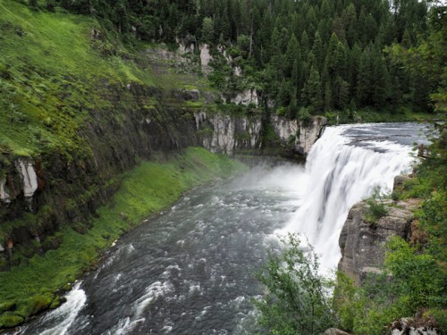 A majestic waterfall cascades into a river, surrounded by lush green trees and rocky cliffs. Mist rises from the water.
