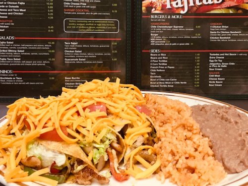 A plate of fajitas topped with cheese, lettuce, and tomatoes, served with rice and refried beans, against a menu backdrop.