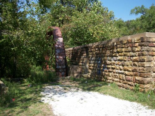 Overgrown path leading to a weathered stone wall and a rusty arch, surrounded by trees and greenery.
