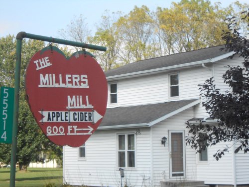 Sign for Miller's Mill, advertising apple cider, with a white house and trees in the background.