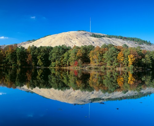 A serene landscape featuring a hill with a tower, surrounded by colorful trees reflecting in a calm lake under a blue sky.