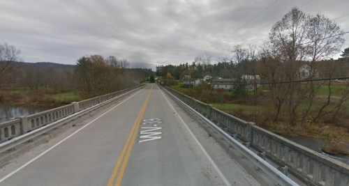 A wide, empty road stretches over a bridge, surrounded by trees and a small town in the background under a cloudy sky.
