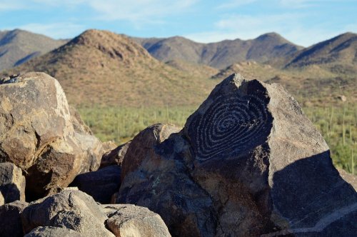A rock with a spiral petroglyph, set against a backdrop of mountains and desert vegetation.