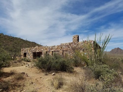 Abandoned stone structure surrounded by desert vegetation and mountains under a cloudy sky.