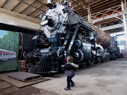 A child walks past a large, vintage steam locomotive inside a train museum.