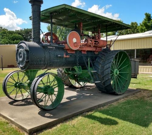 A vintage steam tractor with a green and black exterior, displayed on a concrete base under a blue sky.