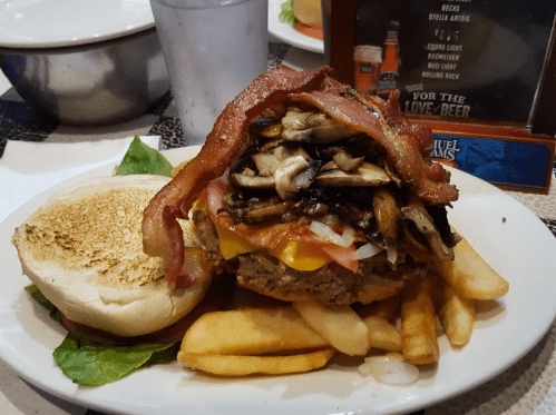 A large burger topped with bacon, mushrooms, and vegetables, served with fries and a toasted bun on a plate.