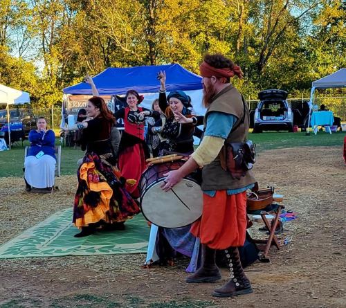 A lively outdoor scene with dancers in colorful costumes performing near a drummer, surrounded by tents and spectators.