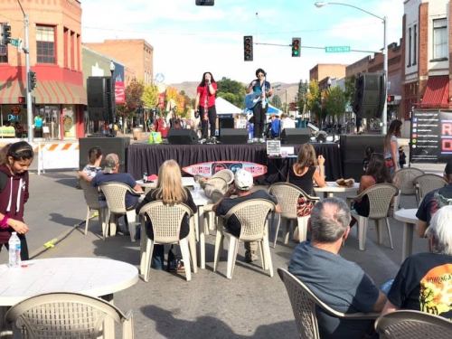 A street performance with two musicians on stage, while an audience sits at tables in front, enjoying the show.