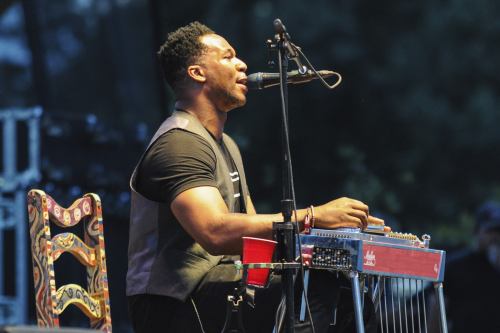 A musician performs on stage, playing a steel guitar and singing into a microphone, with a colorful chair beside him.