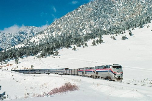 A train travels through a snowy landscape, surrounded by mountains and evergreen trees under a clear blue sky.