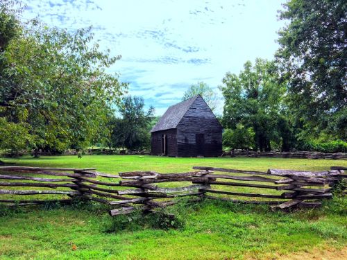A rustic wooden barn surrounded by a split-rail fence, set in a green landscape with trees and a cloudy sky.