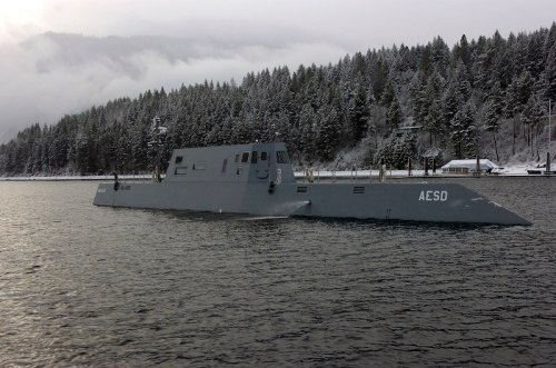 A gray submarine navigates through calm waters, surrounded by snow-covered trees and misty mountains.