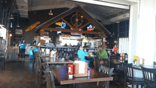 Interior of a casual restaurant with wooden tables, a bar area, and patrons enjoying their meals. Bright neon signs visible.