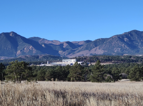 A factory nestled among rolling hills and mountains, with tall grass in the foreground under a clear blue sky.