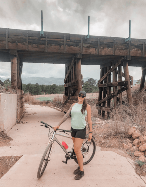 A woman stands with a bicycle near a wooden bridge, surrounded by dry grass and trees under a cloudy sky.