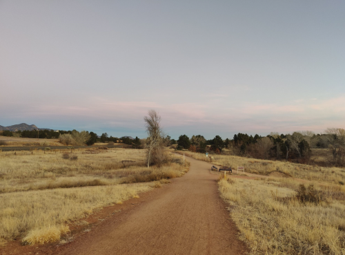 A dirt path winds through a grassy landscape under a pastel sky, with trees and distant mountains in the background.