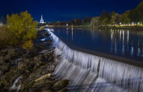 A serene night scene of a waterfall cascading over rocks, with a lit building and trees in the background.