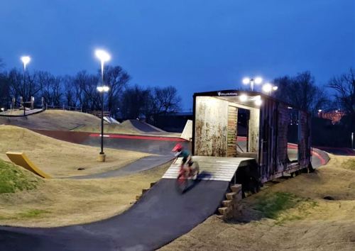 A BMX rider speeds down a ramp at a bike park, illuminated by lights at dusk.