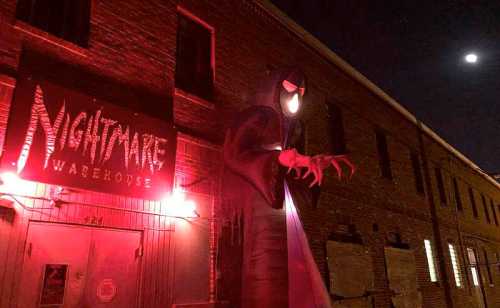 A spooky warehouse entrance with eerie red lighting and a large, menacing figure looming outside under a night sky.