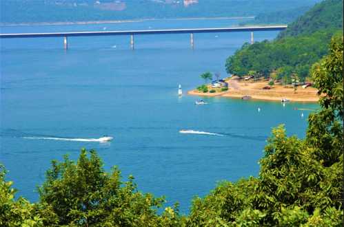 A scenic view of a blue lake with boats, a bridge in the background, and lush green trees along the shore.