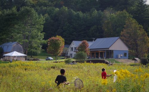 Two people in a field near a house and a tent, surrounded by trees and autumn foliage.