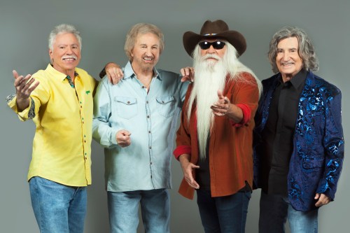 Four men stand together, smiling and posing for the camera, dressed in colorful shirts and jackets against a gray background.