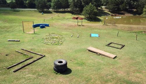 Aerial view of an outdoor training area with various obstacles, including tires, wooden planks, and a small pond nearby.