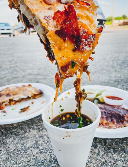 A cheesy, meaty quesadilla being lifted above a cup of sauce, with plates of food in the background on a pavement.