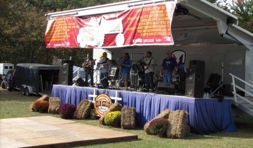 A band performs on an outdoor stage decorated with hay bales and colorful flowers, surrounded by trees.