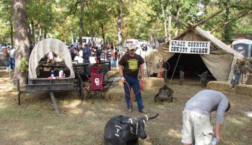 A man leads a toy bull near a tent labeled "Grace Country Cowboy Church" at a lively outdoor event.